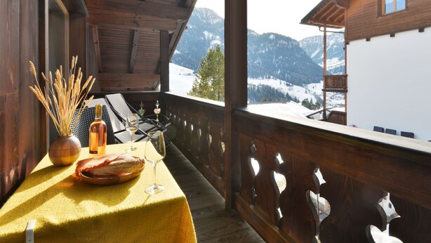 A wooden balcony with a table set for two, featuring a bottle, glasses, and a bread basket. A view of snow-covered mountains is visible through the window. - Apartment Tirol by Interhome (Ortisei St Ulrich)