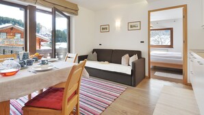 A modern room with a dining table, a sofa, and a bed. There are windows with a view of buildings and trees. - Apartment Tirol by Interhome (Ortisei St Ulrich)