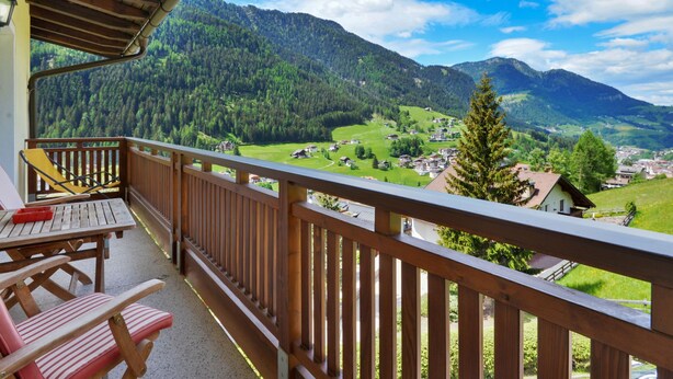 En balkon med trægelænder med udsigt over et bjergrigt landskab med huse og grønt.