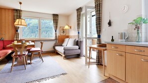 En hyggelig stue med sofa, spisebord og et køkkenområde med træskabe og et vindue med gardiner.