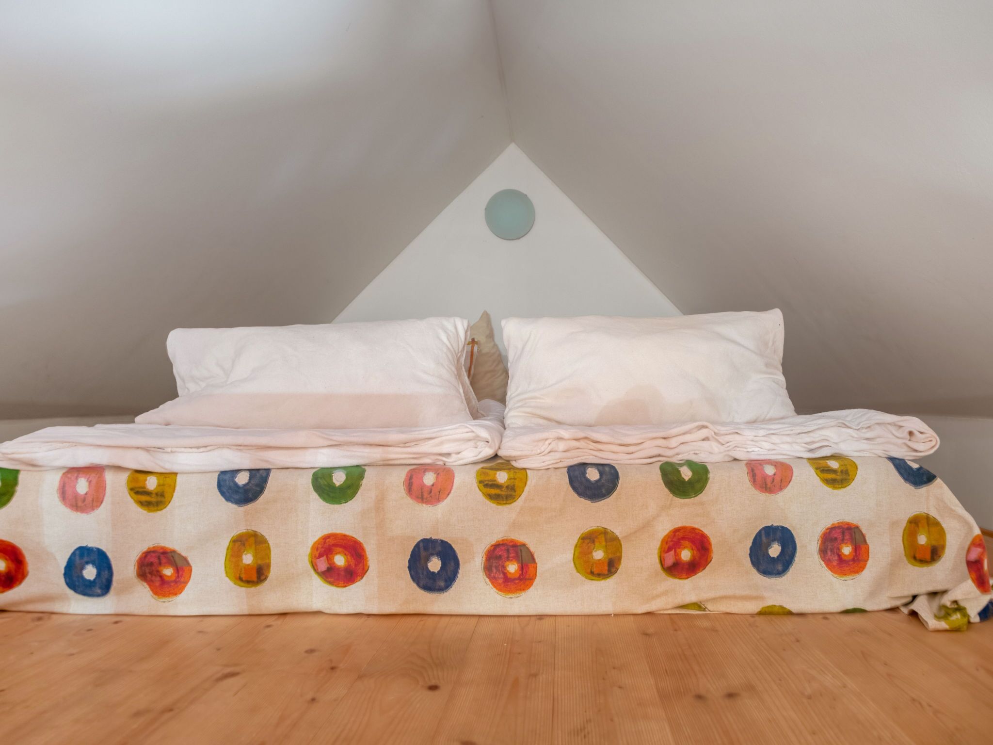 A neatly made bed with a colorful circular pattern on the bedspread, a white pillow, and a white pillow on the headboard.