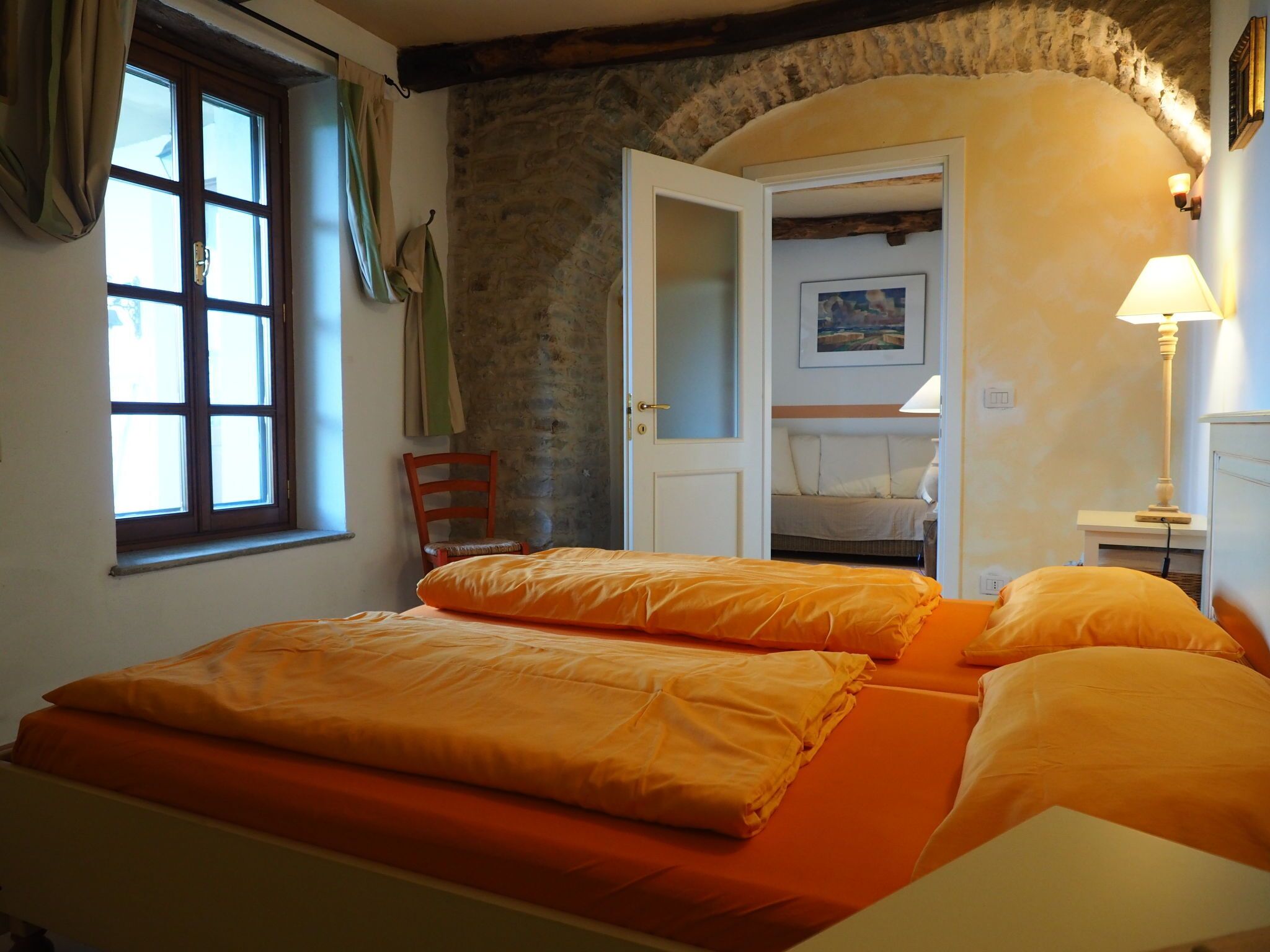 Quarto com cama, cadeira, janela e porta que dá para outro cômodo.
