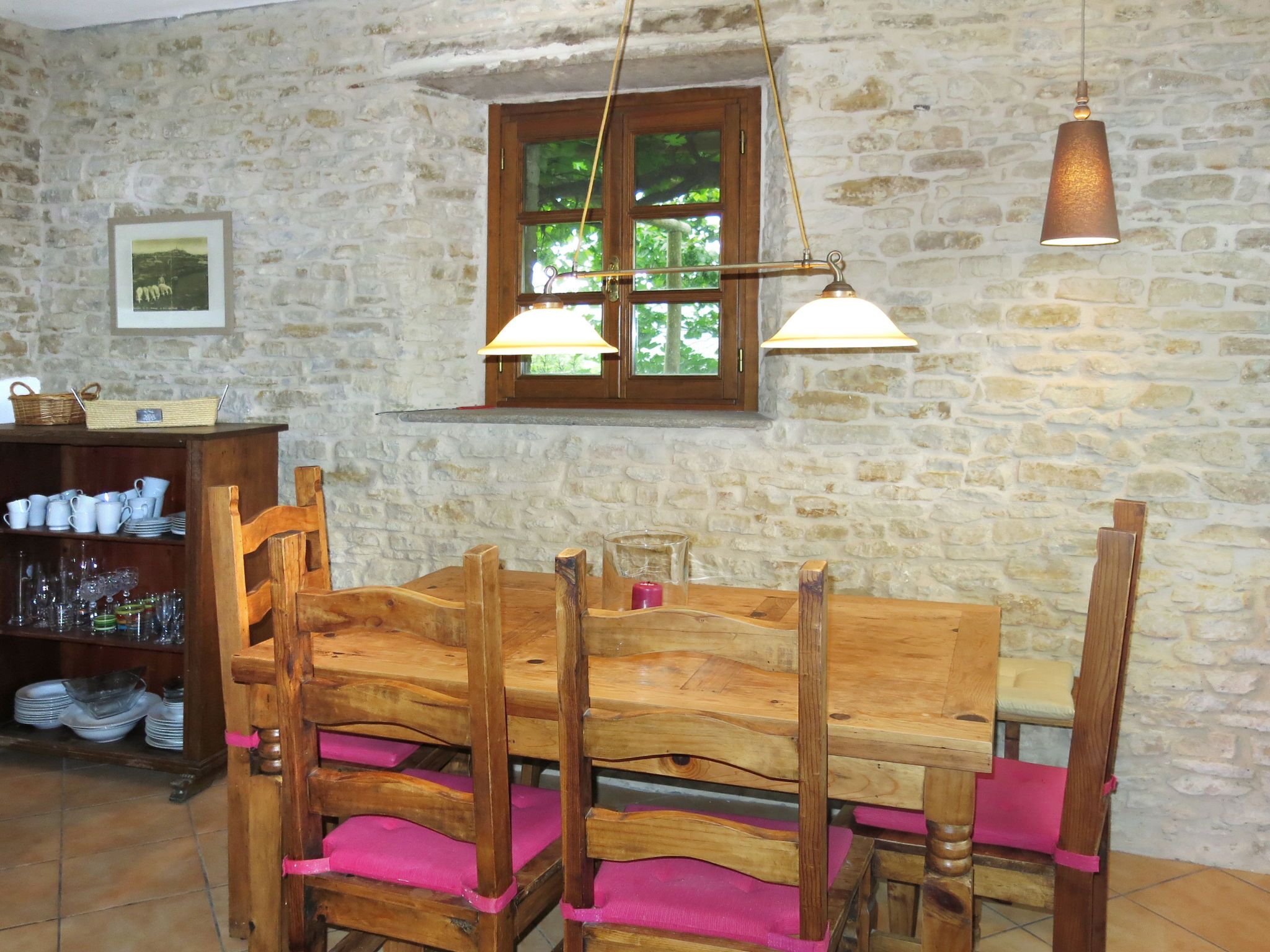 A rustic dining area with a wooden table and chairs, a stone wall, and a window with a view of greenery.