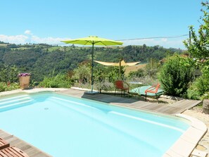 Ein Schwimmbecken mit Holzterrasse, einem grünen Sonnenschirm und Blick auf eine hügelige Landschaft.