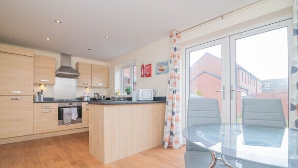 A modern kitchen with wooden cabinets, a central island, and a dining area with a glass table and chairs. - Quiet and Lovely 3-bed house parking and garden (Wolverhampton)