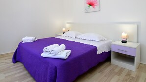 A neatly made bed with a purple bedspread and white pillows, two bedside tables with lamps, and a wall-mounted artwork of a pink flower. - Katarina 4 by Interhome (Šibenik)