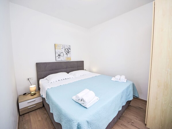 A neatly made bed with a light blue bedspread and white pillows, a bedside table with a lamp, and a wall-mounted artwork above the bed.