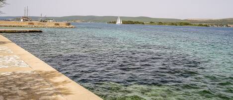 A waterfront view with a clear blue sea, a stone walkway, and distant hills.