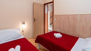 Ein Hotelzimmer mit zwei Betten, jedes mit roter Bettdecke und weißen Kissen. An der Wand befindet sich eine Holztür und ein Lichtschalter.