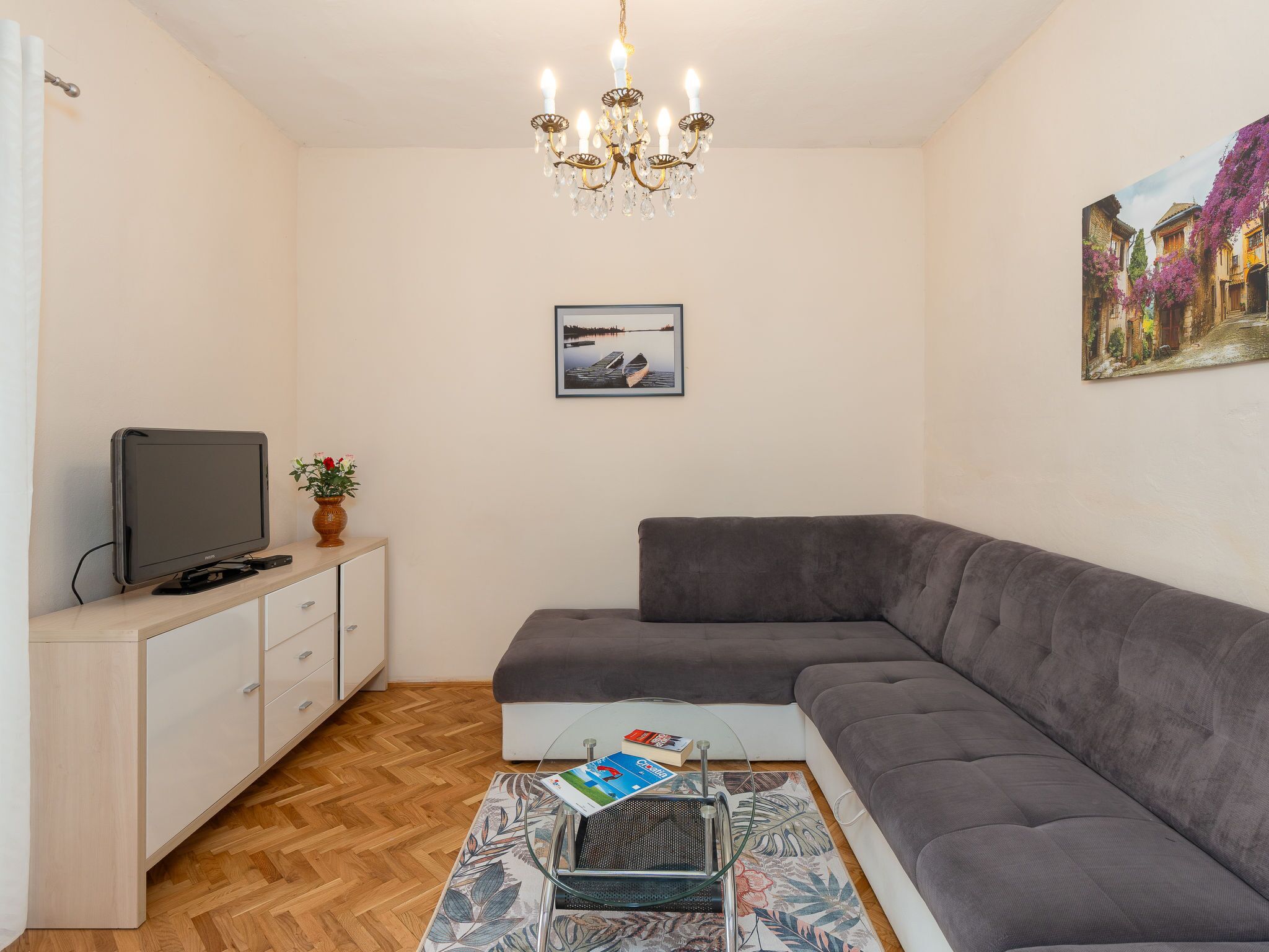 Una sala de estar moderna con un sofá seccional gris, un televisor de pantalla plana sobre un mueble blanco, una mesa de centro de vidrio con un libro, un tapete floral y un cuadro de una escena callejera en la pared.