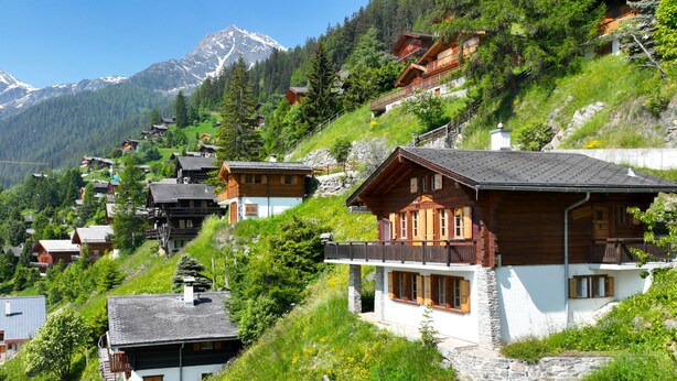 Un borgo montano con case in legno tradizionali, muri in pietra e vista sulle montagne innevate.