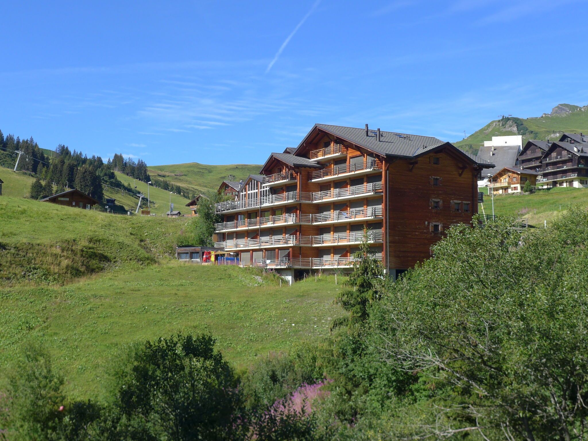 A multi-story wooden building with balconies, set in a green, hilly landscape.