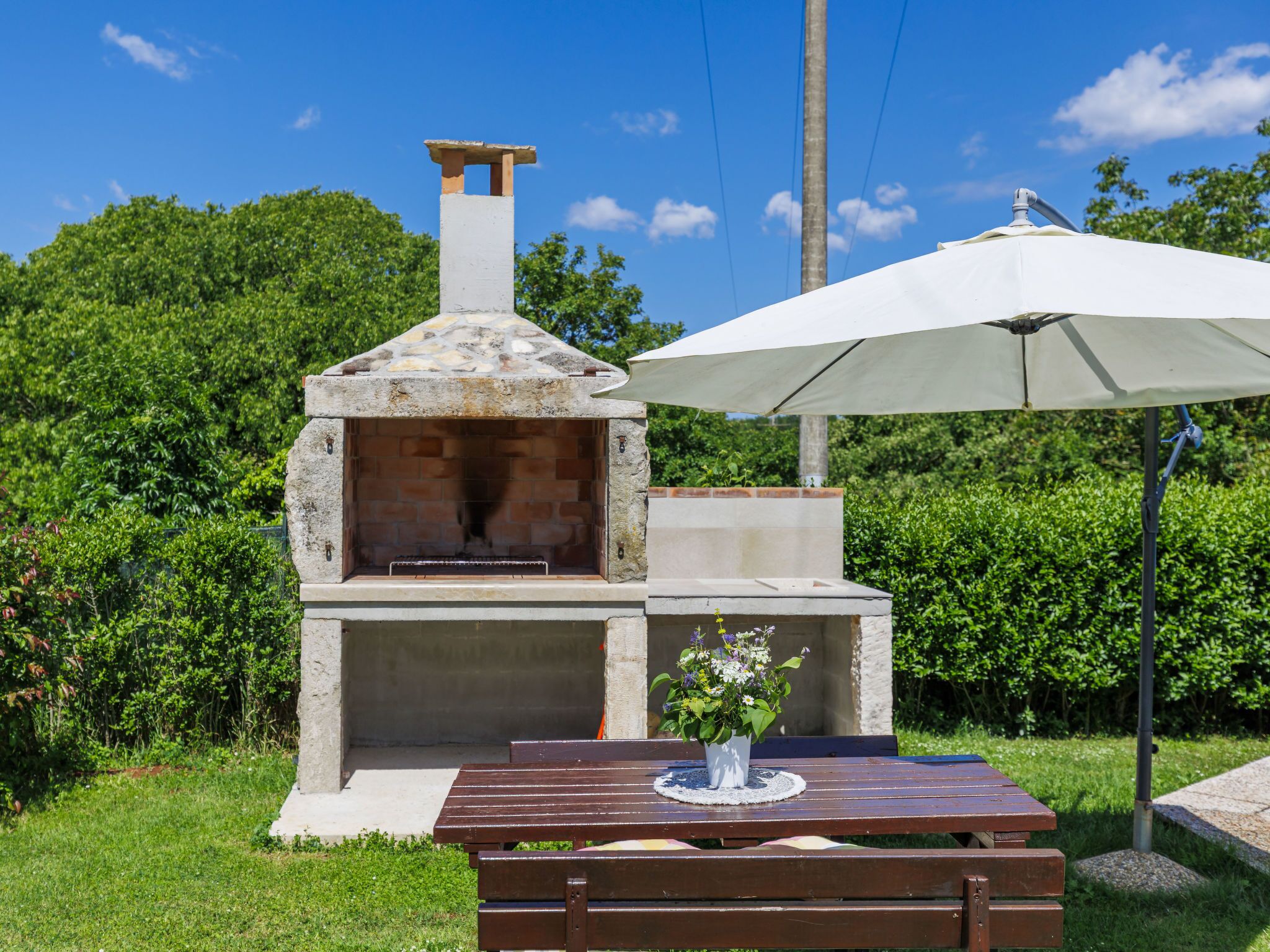 Un horno de pizza al aire libre con chimenea, una mesa de madera con un banco y una sombrilla blanca que da sombra.