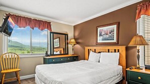A bedroom with a large bed, a wooden nightstand, a chair, a dresser, a mirror, and a window with a view.