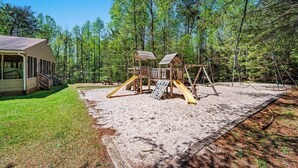 A playground with a wooden swing set and slide, surrounded by a sandy area and a grassy lawn, with a building to the left.