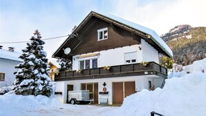 A two-story chalet with a snow-covered roof, a visible garage, and a mountain backdrop.