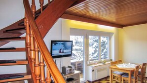 A living room with a wooden staircase, a television, a dining table with chairs, and a view of a snowy landscape through large windows.