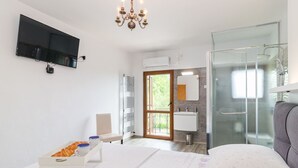 Ein Schlafzimmer mit Bett, Fernseher, einer Kristalllüster, einer Glasdusche und einem Badezimmer mit Waschbecken und Spiegel.