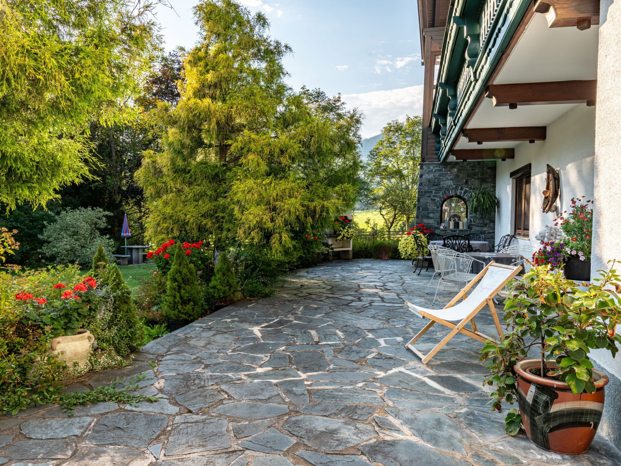 Une terrasse dallée de pierres, dotée de sièges extérieurs, entourée d’une végétation luxuriante et bordée d’un bâtiment en pierre surmonté d’un toit végétalisé.