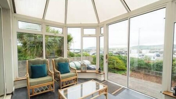 Une véranda meublée de mobilier en osier et dotée d’une table en verre, de grandes fenêtres offrant une vue sur la verdure et un espace de stationnement.