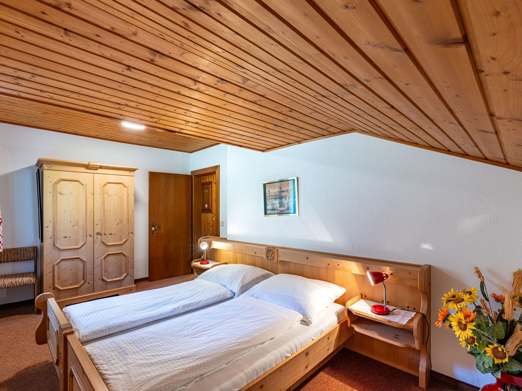 A bedroom with a wooden ceiling, a double bed, a nightstand with a lamp, a wardrobe, and a painting on the wall.