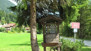 Un cartel de madera con varios indicadores de destino, una señal de tráfico cercana y un exuberante paisaje verde con árboles y casas al fondo.