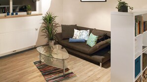 Una sala de estar con un sofá marrón, una mesa de centro de vidrio, un tapete a rayas, una planta en maceta y una estantería.