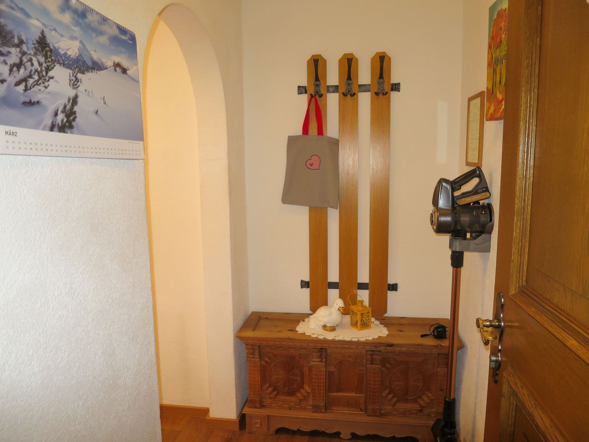 Una habitación con un armario de madera, un calendario, un perchero con abrigos y una cámara sobre un trípode.