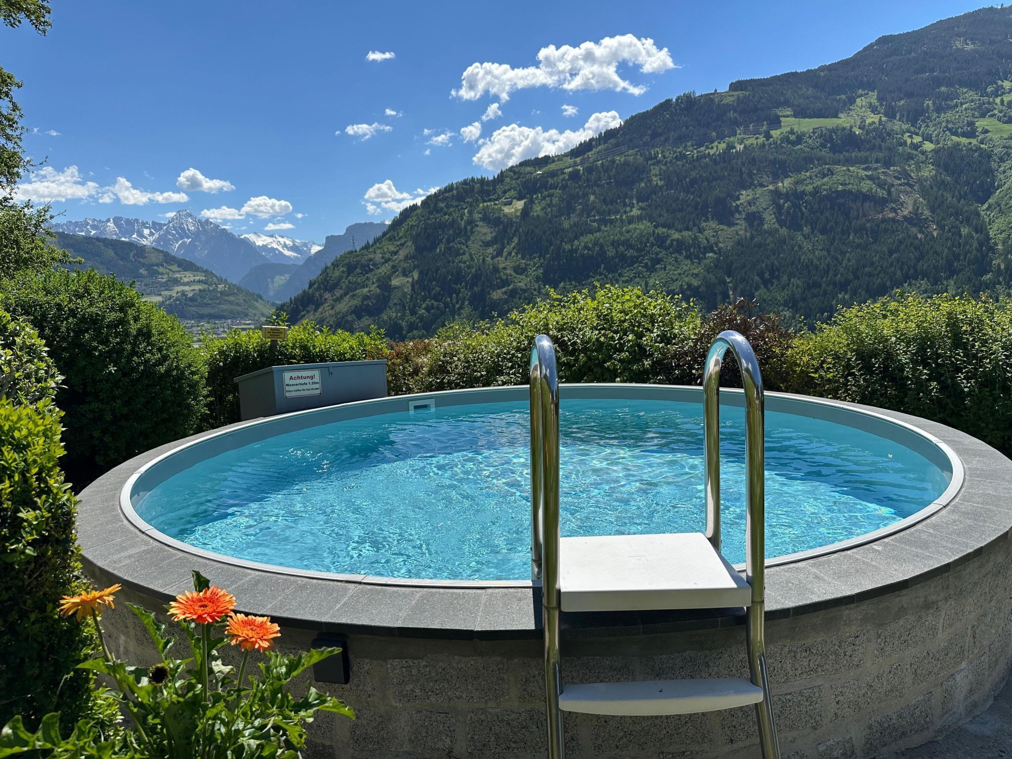 A circular pool with metal ladders, surrounded by greenery and mountains in the background.