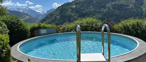 A circular pool with metal ladders, surrounded by greenery and mountains in the background.