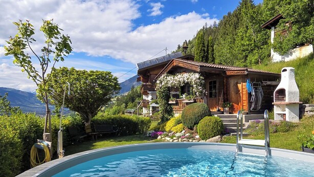 Piscina com deck de madeira, montanhas ao fundo e um edifício rústico com telhado inclinado.