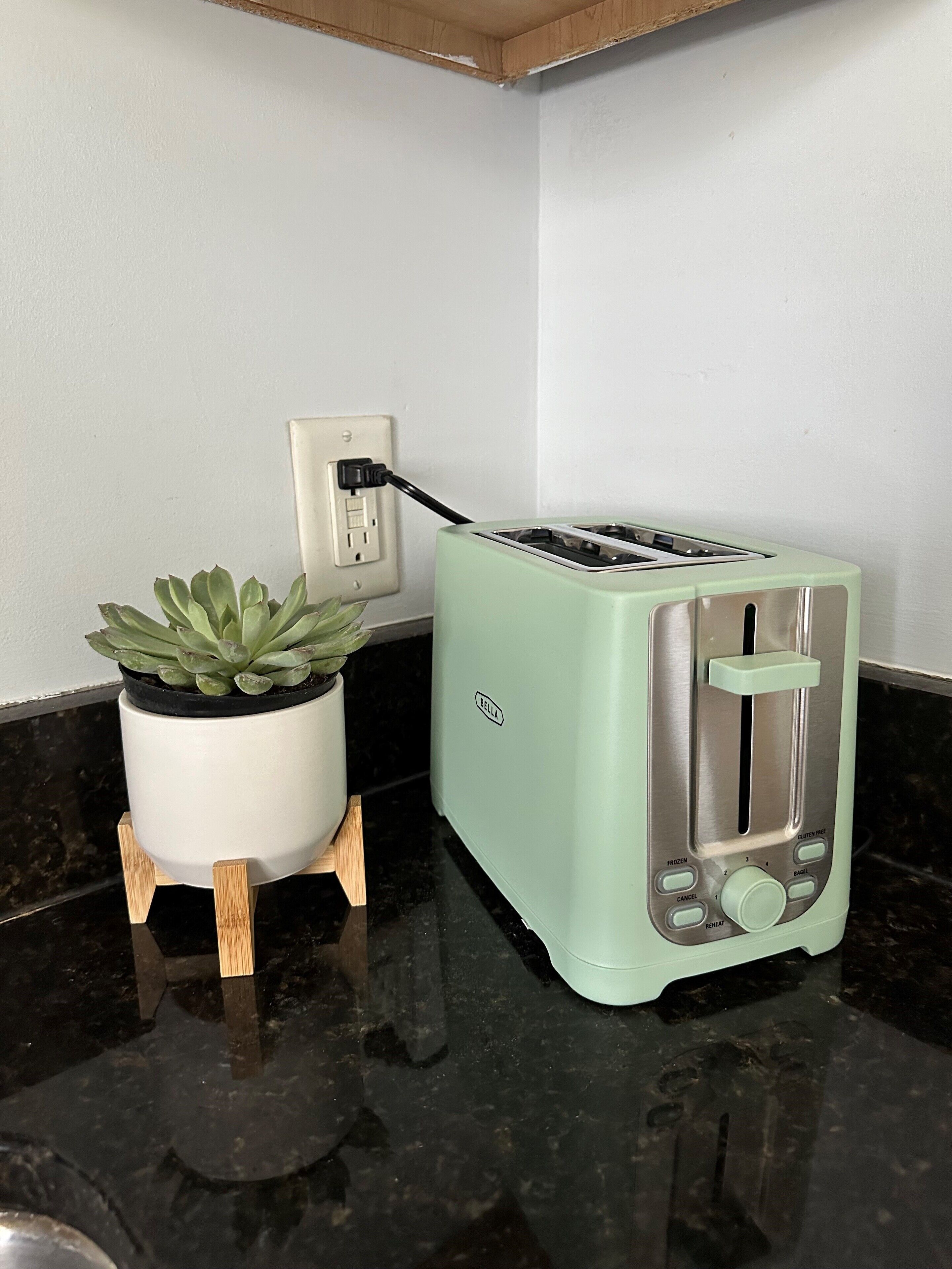 A kitchen with a toaster on a black countertop, a potted plant on a wooden stand, and a white wall with a power outlet.