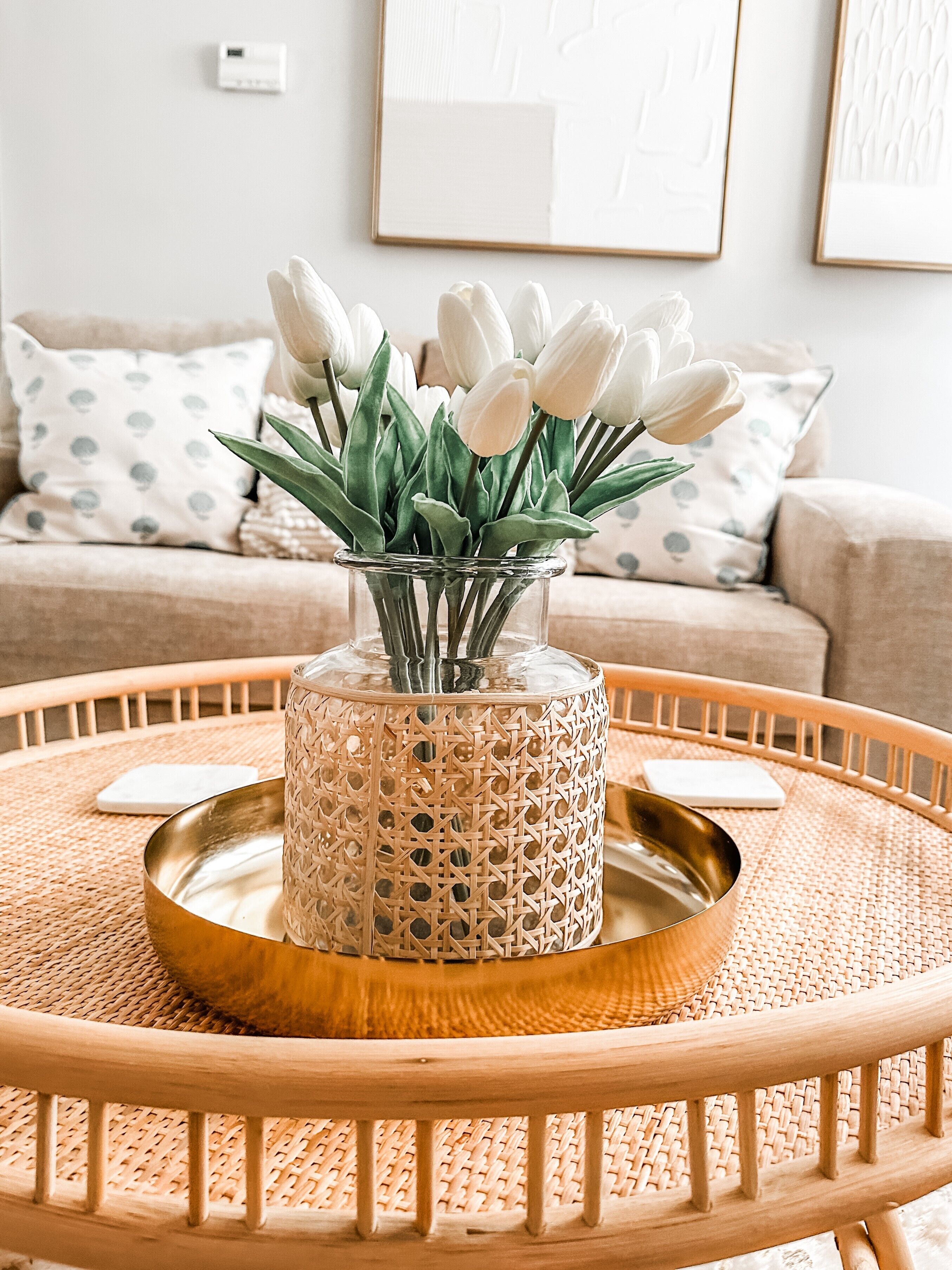 A round wooden table with a woven tray, a decorative glass vase with white tulips, and a beige sofa with patterned cushions in the background.