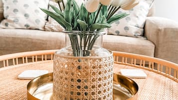 A round wooden table with a woven tray, a decorative glass vase with white tulips, and a beige sofa with patterned cushions in the background.