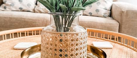 A round wooden table with a woven tray, a decorative glass vase with white tulips, and a beige sofa with patterned cushions in the background.