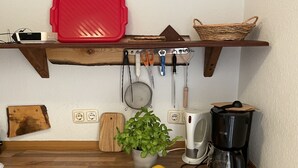 A kitchen with a wooden shelf holding a red tray, a basket, and a Philips device. Below, there are two power outlets, a wooden cutting board, a potted basil plant, two lemons, a coffee maker, and a trash can. - Old Hüsung (MUZ321) by Interhome (Waren)