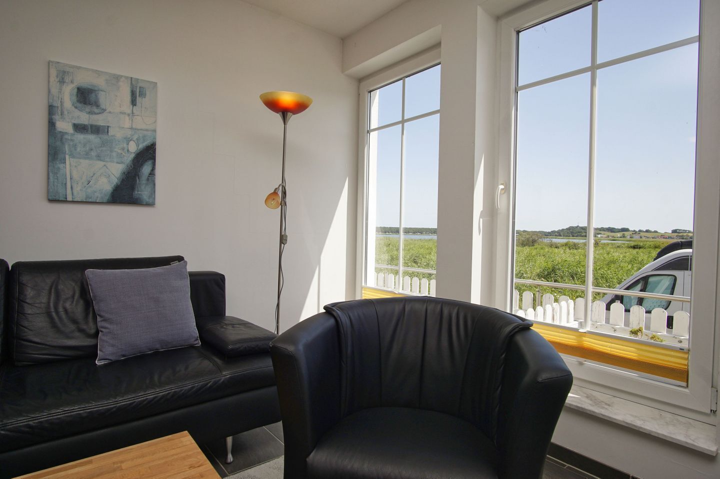 A room with a black leather sofa, a single chair, a floor lamp, and a large window with a view of greenery and a fence.