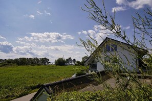 A house with a sloped roof and large windows, surrounded by a green field and trees.