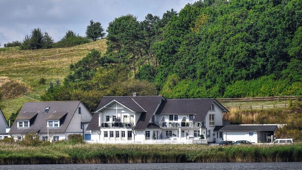 A coastal property with multiple houses, a dock, and a hill with dense greenery in the background. - Chic vacation apartment with sea view - rügenreif Holiday apartment no. 02 - rügenreif (Mönchgut)