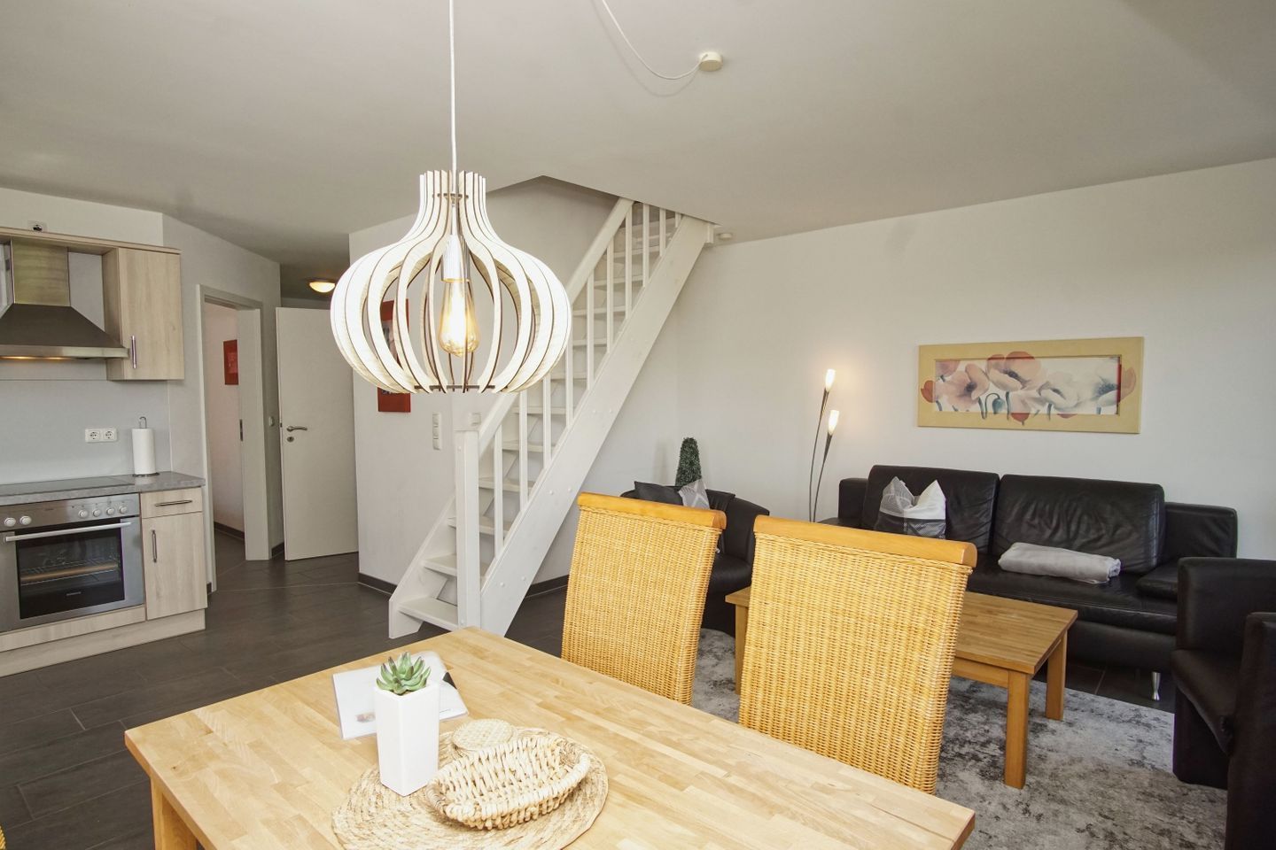 Eine moderne Küche mit integrierten Geräten, ein Essbereich mit Holztisch und Korbstühlen, eine Treppe und ein Wohnzimmer mit Sofa und einem Gemälde.