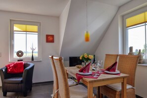 A dining area with a wooden table set for four, surrounded by wicker chairs, a black sofa, and a red throw pillow. - Chic vacation apartment with sea view - rügenreif Holiday apartment no. 02 - rügenreif (Mönchgut)