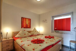 Ein Schlafzimmer mit einem Bett, zwei Nachttischen, einem Fenster mit roten Jalousien, einem Wandbild und einer kleinen Topfpflanze.
