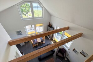 A high-ceilinged room with a wooden beam structure, a dining area with a table and chairs, and large windows offering a view of the outdoors.