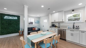 A modern kitchen with white cabinets, a wooden dining table with blue chairs, and a neon sign on the wall. - GAME ROOM-Downtown Remodeled Single Family House (Ocean City)