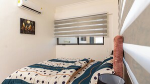 A bedroom with a bed, a window with blinds, an air conditioner, and a painting on the wall.