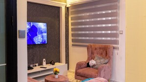 Een woonkamer met een roze bank, een televisie, een raam met jaloezieën en een plafondventilator.