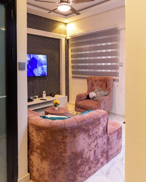 Een woonkamer met een roze bank, een televisie, een raam met jaloezieën en een plafondventilator.