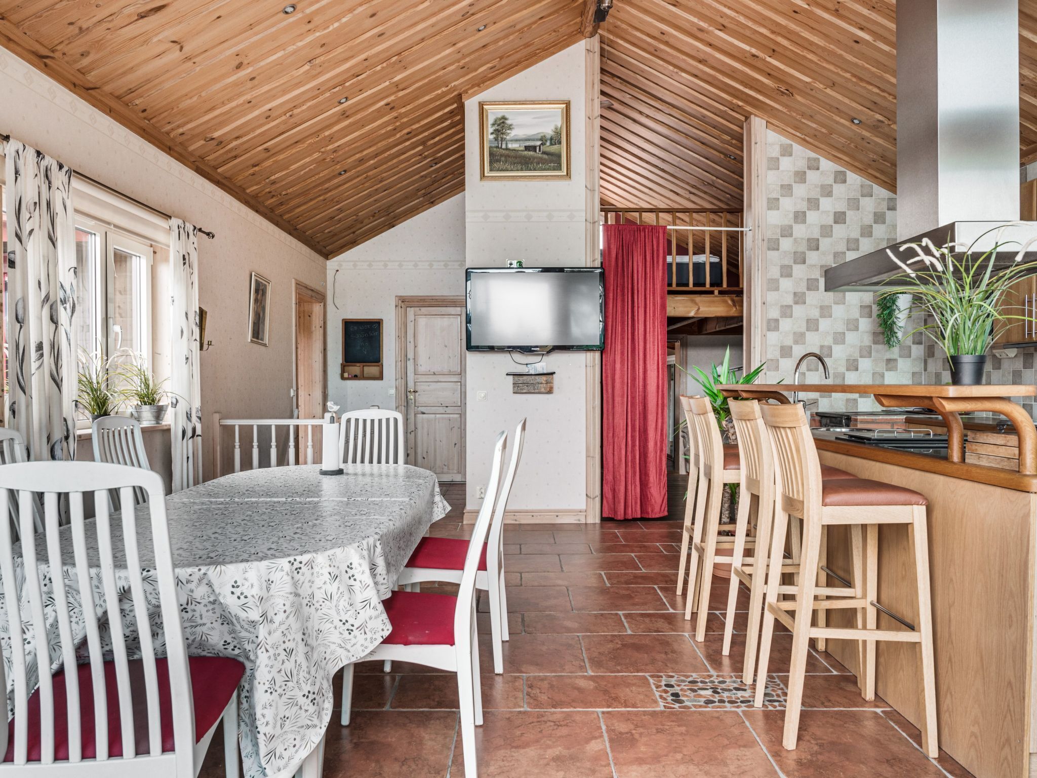 A cozy, well-lit room with a wooden ceiling, a dining table with chairs, a kitchen area with a sink and counter, and a television mounted on the wall.