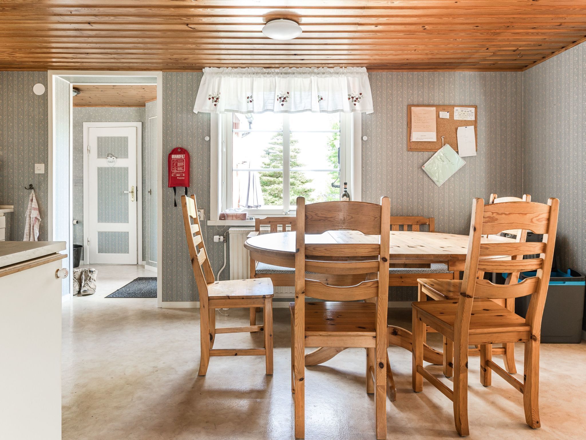 Ein gemütliches Zimmer mit Holzboden, einem runden Holztisch mit Stühlen, einem Fenster mit weißem Vorhang und einer Pinnwand mit Papieren.