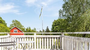 A wooden deck with a white railing, a red building in the background, and a flagpole with a flag. - Gåragöl by Interhome (Olofström)